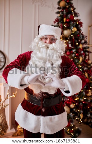 Santa Claus forming shape of heart with her fingers near the fireplace and christmas tree with gifts. New year and Merry Christmas , happy holidays concept