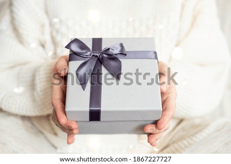 The girl's hands in a cozy white sweater are holding a Christmas present in a gray box with warm Christmas lights.