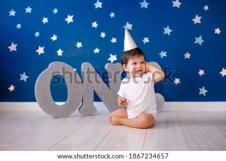 Baby boy of one year old wearing a white body and a festive paper hat sits on the floor on a blue background with stars and big silver letters one.