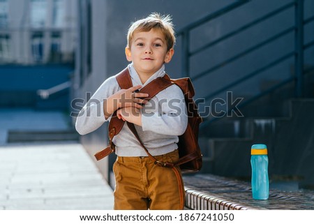 Cute blond little boy going first time to school. Pupil of primary school holding bottle with backpack at sunny day outdoor.