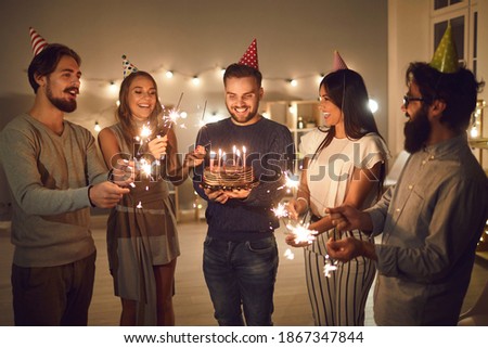 Birthday celebration at home concept. Group of young cheerful friends in decorative caps congratulating their man friend with cake in hands with birthday and holding festive sparklers in hands