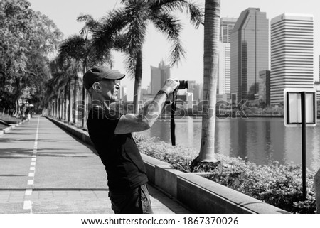 Profile view of senior tourist man wearing cap while taking pictures at peaceful park in Bangkok Thailand