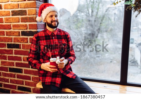 Attractive christmas man wearing santa claus hat sits on windowsill and looks out the window enjoying Merry Christmas at home, Merry Christmas and Happy New Year