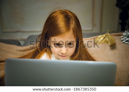 Comfortable. Happy caucasian little girl during video call with laptop and home devices, looks dreamful and happy. Talking to Santa before New Year's eve, her family, watching cartoons, typing text