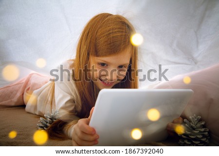 Miracle. Happy caucasian little girl during video call with laptop and home devices, looks dreamful and happy. Talking to Santa before New Year's eve, her family, watching cartoons, typing text. Bokeh
