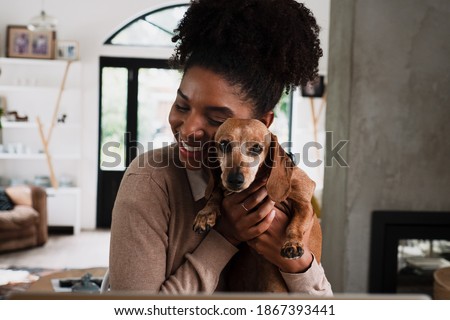 African American female smiling while snuggling cute puppy sitting at desk in modern kitchen. Royalty-Free Stock Photo #1867393441