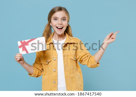 Excited little blonde kid girl 12-13 years old in yellow jacket posing isolated on blue background. Childhood lifestyle concept. Mock up copy space. Hold gift certificate, pointing index finger aside