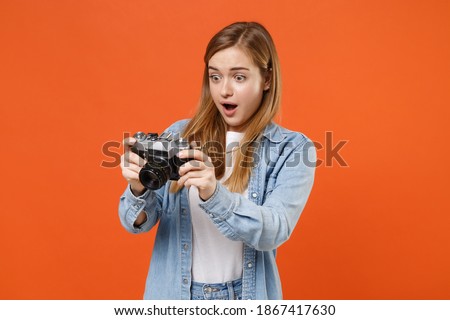 Shocked young woman 20s in casual denim clothes posing isolated on orange background studio portrait. People sincere emotions lifestyle concept. Mock up copy space. Hold retro vintage photo camera