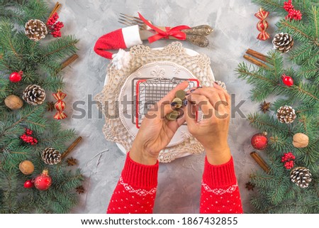 Christmas gray grunge background, fir twigs, berries, snowflakes, baubles, top view. New Year table setting menu decoration. Tiny shopping basket, hands in red pullover count coins, economy, poorness