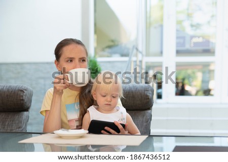Young woman in cafe drinks a coffee while child in her arms looks at phone. Modern business mom.