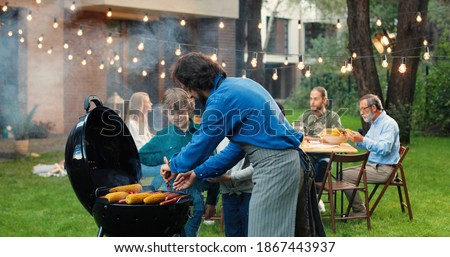 Father cooking food on barbecue for weekend lunch and giving hot meat and corn to small cheerful kids. Family at picnic on back yard. Little son and daughter at dad waiting as man preparing eating. Royalty-Free Stock Photo #1867443937