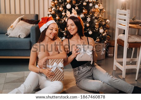 Two beautiful young girls with Christmas gifts in their hands are sitting near the Christmas tree in the house.friends with Christmas dresses and wrapped gifts