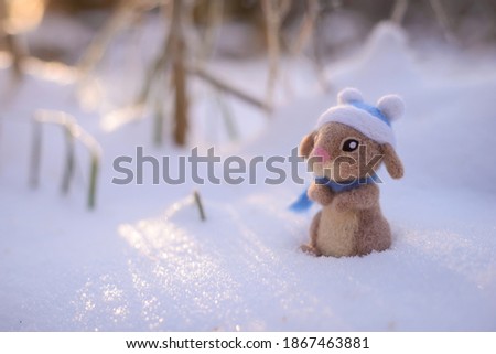Toy wool felted cute rabbit, hare in a blue hat and scarf stands in the snow on a sunny winter day