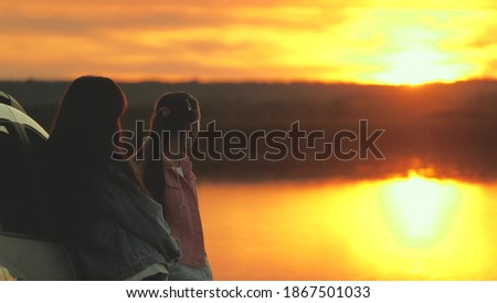 Happy mothers and daughter hug, travel, stand next to car and admire beautiful sunset on beach. Free women tourists by car, admiring sunrise, river. Family travelers, tourists. Family travel by car.