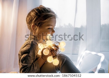 Happy little girl looks in the mirror while sitting by the window. Magical portrait of a little girl in a New Year's style.