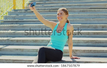 Happy smiling young fitness woman taking selfie with smartphone in the city park