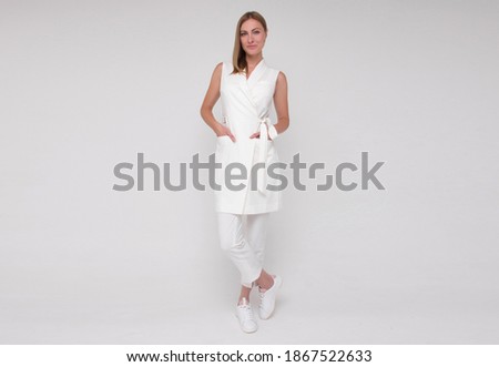 young stylish nurse in white medical costume and white sneakers is standing straight with hands in her pockets and smiling on white wall background. medical concept. free space