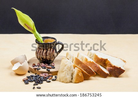 Still life composition with wooden kitchen cutting board, beans, egg shell, bread and ceramic pot with Arum flower on dark background. Image has grain texture visible on its maximum size