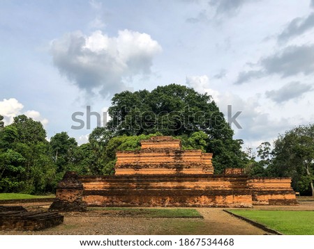 A photo of Candi Muaro Jambi, a complex of ancient Buddhist temples in Indonesia. The photo shows the red brick structures surrounded by green trees and grass. 