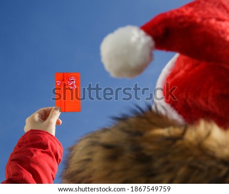 Close-up of a female hand holding a gift card with a picture of sweets against a blue sky. Wearing a Santa hat and a red jacket. gift or discount concept. selective focus