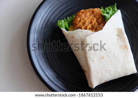 Delicious tortilla wrap with fried chicken meat and green coral lettuce or salad on black plate.