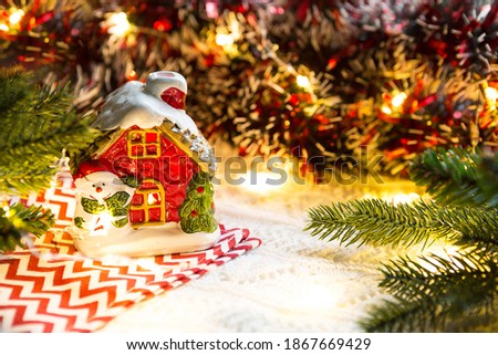 Christmas toy-a house with glowing Windows and a snowman in a cozy setting. New Year, napkin with a zigzag, in the lights of garlands. Holiday decor, comfort, knitted blanket, spruce branches