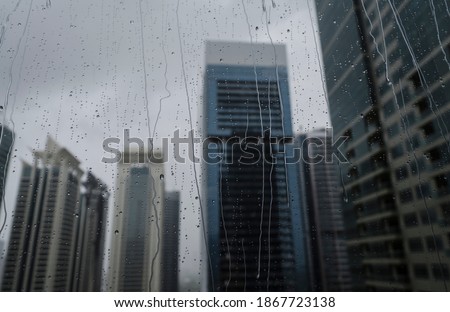 A blur view of office towers through wet window glass pane. Abstract photo of modern building during rainy season.