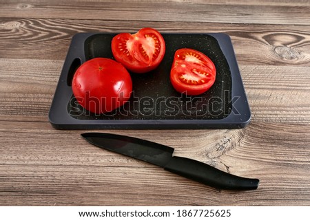 One whole tomato and another, cut in half, lie on a cutting Board on the wooden surface of the table. Next to it is a black knife. Image with selective focus.