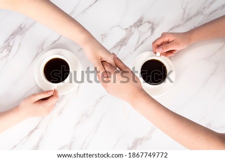 Close up of the hands of a woman sitting face to face, reaching out, holding a coffee cup with one hand and holding each other's hands with one hand