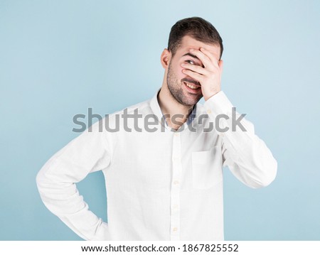 Emotive handsome and joyful guy in white shirt covering eyes with palms and peeking through fingers with one eye smiling over blue background