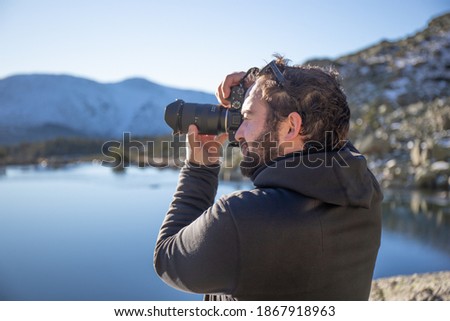 A man taking pictures in black sweatshirt in a blue lake with snowy mountains