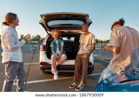 Hanging out. Four young casually dressed friends smiling and speaking to each other having a meeting outside on a parking site near their cars with their redhead girl sitting inside an opened trunk