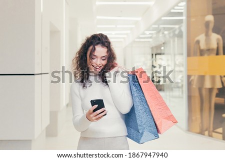 Beautiful brunette girl with curly hair. In a modern clothing store, walks through the supermarket, and holds in his hands colored gift bags with goods, talking on the phone and taking photos