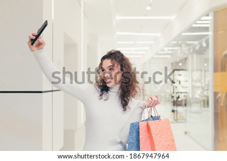 Beautiful brunette girl with curly hair. In a modern clothing store, walks through the supermarket, and holds in his hands colored gift bags with goods, talking on the phone and taking photos