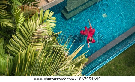 Aerial view of young brunette woman swimming in red dress in the transparent turquoise pool water. Top view of slim lady relaxing on her holidays.