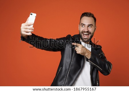 Cheerful young bearded man 20s wearing basic white t-shirt black leather jacket doing selfie shot pointing index finger on mobile phone isolated on bright orange colour background studio portrait