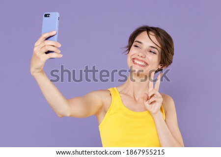 Smiling young brunette woman in yellow casual tank top posing isolated on pastel violet background studio portrait. People lifestyle concept. Doing selfie shot on mobile phone, showing victory sign