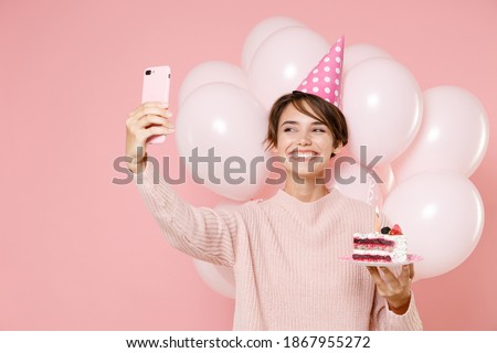 Smiling young woman in casual sweater birthday hat celebrating on pastel pink background with air baloons. Birthday holiday party, people emotions concept. Hold cake doing selfie shot on mobile phone