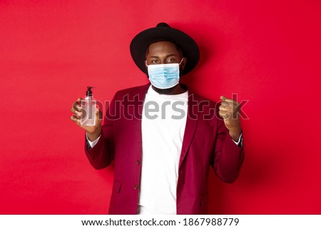 Covid-19, quarantine and holidays concept. Cheerful black man rejoicing of winning, achieve goal, showing antiseptic, wearing medical mask, standing against red background