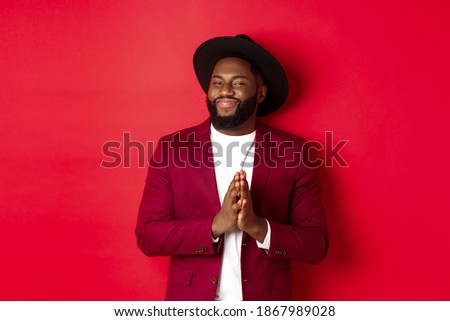 Handsome successful Black man relish profit, rub hands and smiling satisfied, standing against red background in party outfit