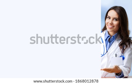 Happy smiling young female doctor in white coat showing blank signboard with copy space empty area for some text, at office. Young brunette beautiful woman - medical, clinic healthcare concept picture