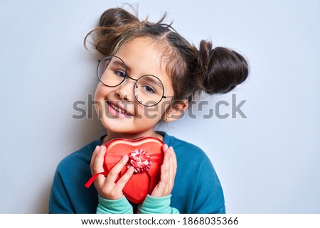 Smiling caucasian little girl with funny hairstyle and glasses holding red box heart gift and looking at camera isolated on white background with blank space. Christmas and New Year present