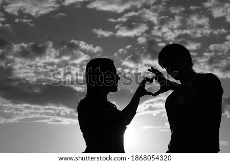 Silhouette of a young girl and her boyfriend with hearts made of fingers against the blue sky with clouds at sunset. Silhouette of the heart.Lovers in nature. Lovers silhouette. Black and white