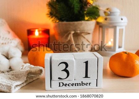 New Year composition with wooden calendar date December 31. Beautiful cozy home decor composition with candles, lantern, Christmas tree, calendar, knitted sweater and tangerines. Selective focus