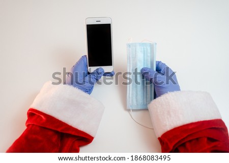 Santa Claus hands holding smart phone and a medical mask over white table background. Online greetings, ordering services for Christmas and new year.
