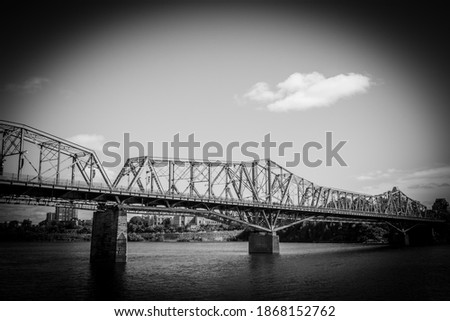 A black and white vignette photo of an old, long, metal bridge stretching across a wide river.