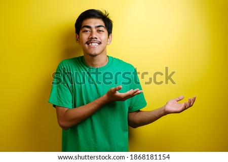 Smiling handsome Asian man pointing finger to empty open hand isolated on yellow background