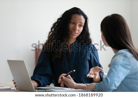 Concentrated multiethnic female employees sit at desk in office work together using computer. Focused women colleagues coworkers cooperate on laptop, brainstorm discuss business ideas at meeting. Royalty-Free Stock Photo #1868189920