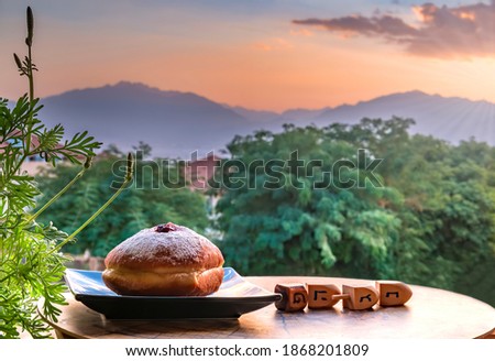 Sweet donates on saucer with wooden dreidels for Hanukkah Holiday - Hebrew letters on  side of dreidel means - Great Miracle Happened Here. Blurred morning sky and mountains, focus on donut

