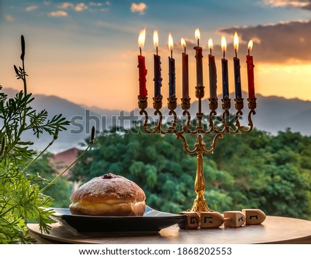 Burning candles, menorah and sweet donates on plate with wooden dreidels for Hanukkah Holiday - Hebrew letters on  side of dreidel means - Great Miracle Happened Here. Blurred morning sky and mountain